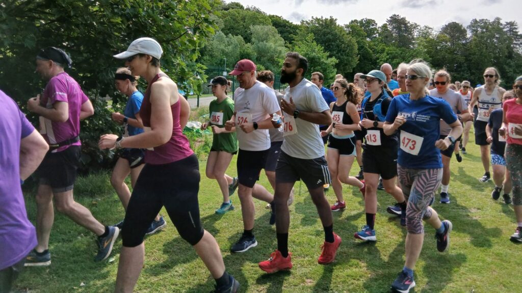 Lots of runners of different ages and gender running past with trees in background.