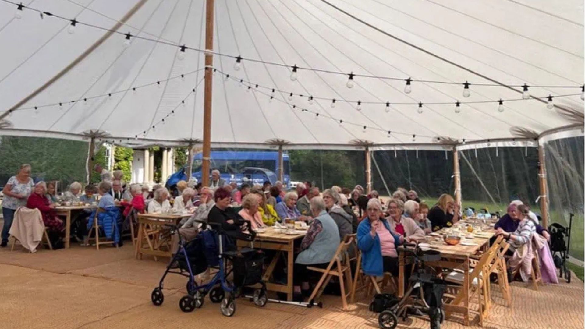 Lots of elderly people sitting on chairs around wooden tables, under a white canopy.